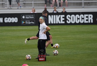 A person in sports attire stands on a soccer field, surrounded by several soccer balls and a carrier with bottles. The field is bordered by advertisements for Nike Soccer and app stores. Several people sit in bleachers in the background.