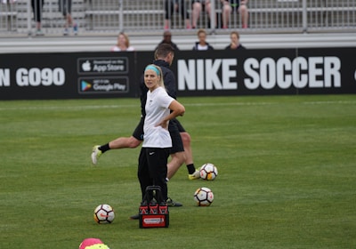 A person in sports attire stands on a soccer field, surrounded by several soccer balls and a carrier with bottles. The field is bordered by advertisements for Nike Soccer and app stores. Several people sit in bleachers in the background.